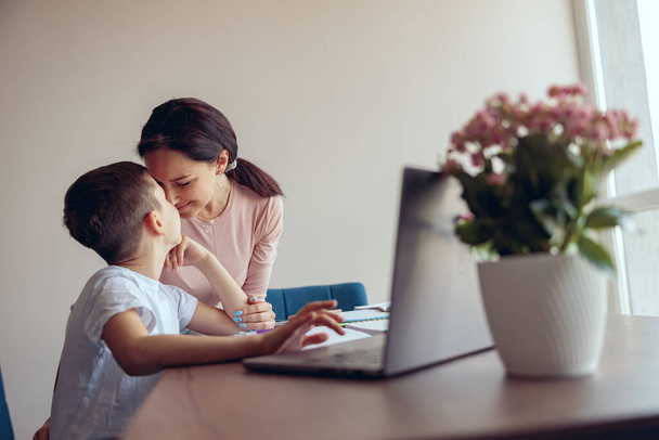 Glückliche Mutter und kleiner Teenager-Sohn, die Nase an Nase zu Hause kuscheln, während sie online lernen. Hausaufgaben machen. - Foto, Bild