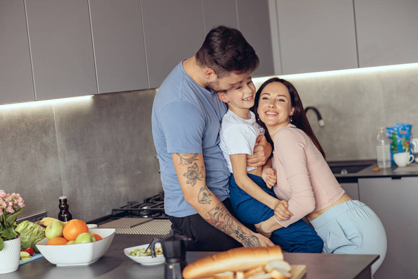 Familia alegre caucásica con un niño pequeño abrazándose en la cocina por la mañana. Mamá y papá sosteniendo hijo. - Foto, Imagen