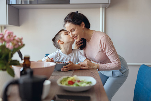 Portrait of cute small boy having breakfast and kissing his mom at table in kitchen. Eating cereals. - Fotografie, Obrázek