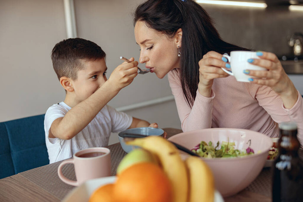 Kleine schattige zoon geeft om zijn ontbijt te proberen aan moeder die koffie drinkt in de ochtend. Ontbijt. - Foto, afbeelding