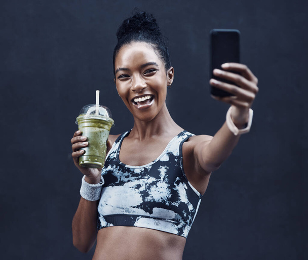 One happy young mixed race woman drinking a healthy green detox smoothie and taking selfies on phone while exercising against dark background. Female athlete sipping on fresh nutritious fruit juice in - Photo, image