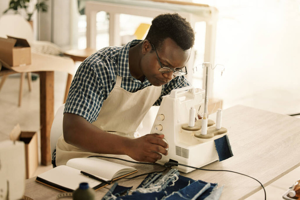 Afrykański amerykański krawiec szyjący dżinsy. Młody projektant korzystający z maszyny do szycia. Poważny krawiec szyjący kawałek tkaniny. Skupiona krawcowa pracuje nad swoim materiałem. - Zdjęcie, obraz