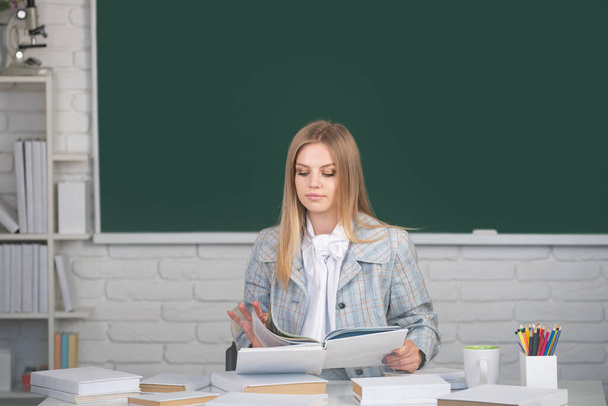 Portrait of young female college student studying in classroom on class with blackboard background. Cute student girl reading book on blackboard background with copy space - Zdjęcie, obraz
