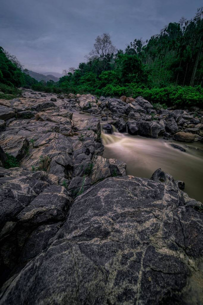 A vertical shot of small stream flowing through rocks and lush green trees in long exposure - Foto, imagen
