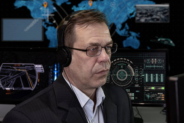 A special agent with headphones looks at the monitor against the background of the world map. Concept: missile launch, surveillance of people, military analytical center, military operations. - Foto, Imagen