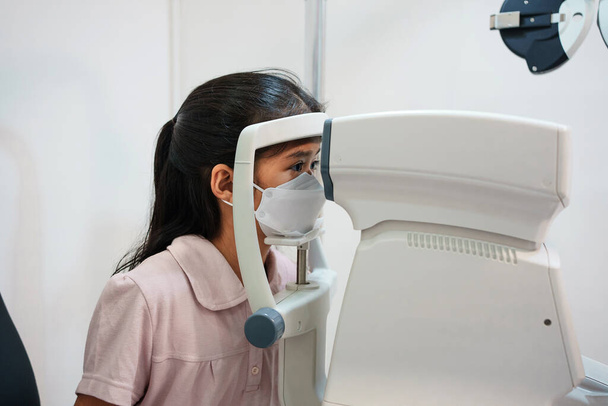 Szemész egy ázsiai lány szemeit vizsgálja egy klinikán. Védőmaszkot viselnek.. - Fotó, kép