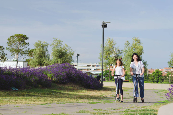 Happy family lifestyle and holiday concept. Mother, little girl, riding scooters, walking in old city, street. Laughing on a summer sunny day. Having fun - 写真・画像