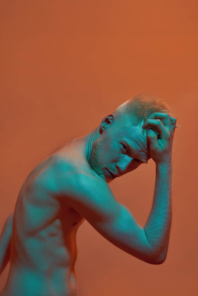 Serious albino caucasian guy touching his head and looking at camera. Young modern bearded blonde man of generation Z with naked torso. Isolated on orange background. Studio shoot. Copy space - 写真・画像