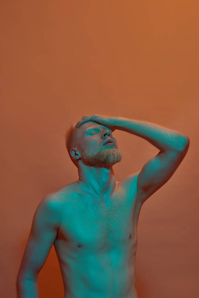 Albino caucasian guy with closed eyes touching his head with hand. Young bearded blonde man of zoomer generation with naked torso. Isolated on orange background. Studio shoot. Copy space - Фото, изображение