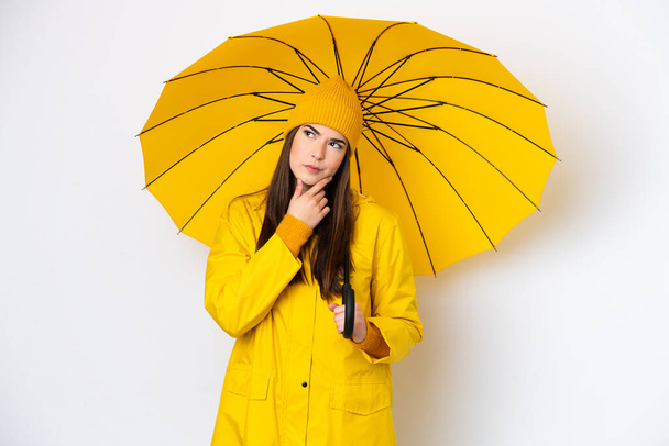 Young Brazilian woman with rainproof coat and umbrella isolated on white background having doubts - Fotografie, Obrázek