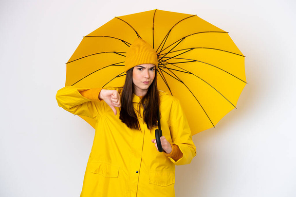 若いですブラジルの女性でレインプルーフコートと傘は、負の式で親指を下に示す白い背景に隔離 - 写真・画像