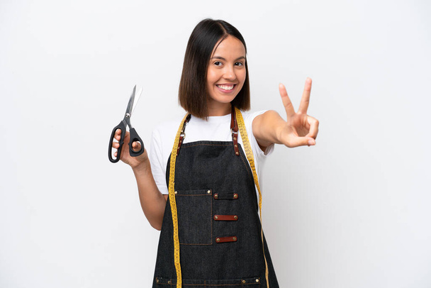 Young seamstress woman isolated on white background smiling and showing victory sign - Foto, imagen