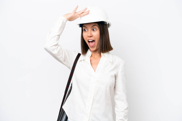 Young architect woman with helmet and holding blueprints isolated on white background doing surprise gesture while looking to the side - Foto, Imagem