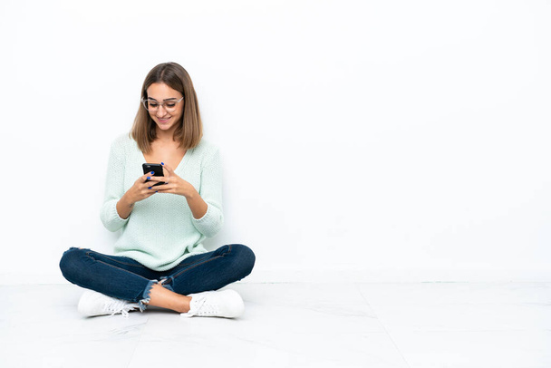 Young caucasian woman sitting on the floor isolated on white background sending a message with the mobile - Фото, зображення