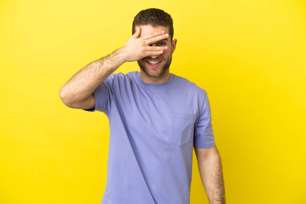 Schöner blonder Mann über isoliertem gelben Hintergrund, der die Augen mit den Händen bedeckt und lächelt - Foto, Bild