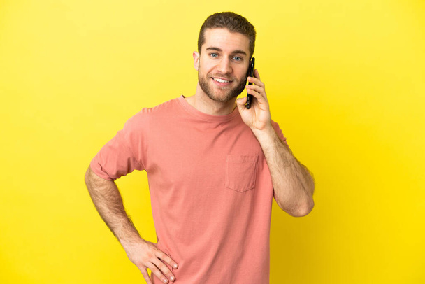 bel homme blond utilisant téléphone mobile sur fond isolé posant avec les bras à la hanche et souriant - Photo, image