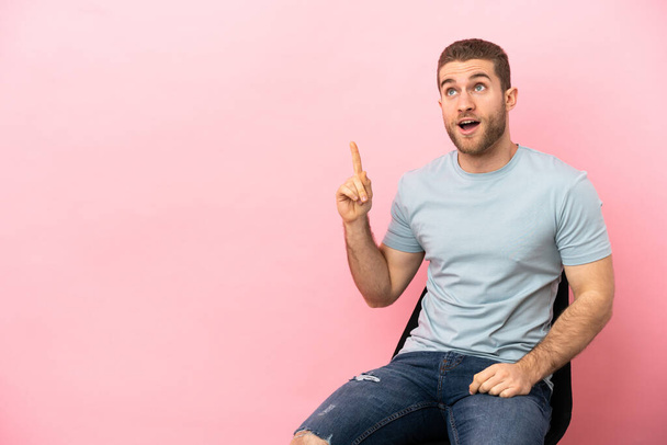 Young man sitting on a chair over isolated pink background thinking an idea pointing the finger up - Foto, Bild