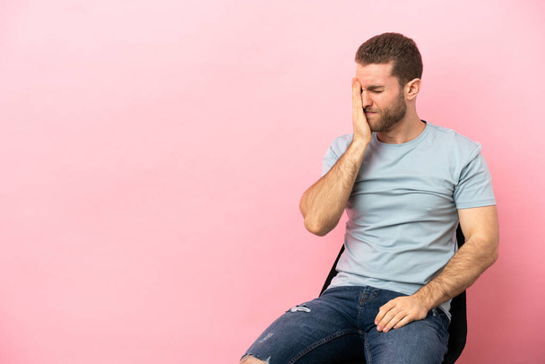 Jovem sentado em uma cadeira sobre fundo rosa isolado com dor de cabeça - Foto, Imagem