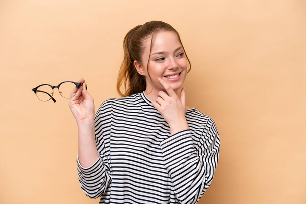 Menina caucasiana jovem com óculos isolados no fundo bege pensando uma ideia e olhando lado - Foto, Imagem