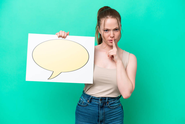 Young caucasian woman isolated on green background holding a placard with speech bubble icon doing silence gesture - Zdjęcie, obraz