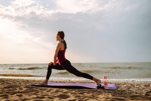Beautiful woman doing yoga near the ocean barefoot. Young woman in sportswear doing fitness exercise on the beach. Calmness and balance, a healthy lifestyle. - Foto, Imagem