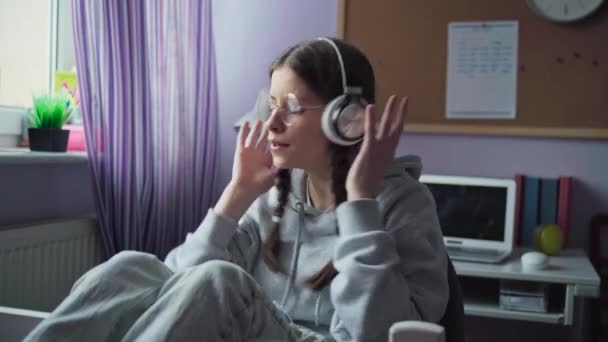Young girl gestures hard and fast and sings while listening to music on wireless headphones. Teenage girl wearing glasses and braids. - Felvétel, videó