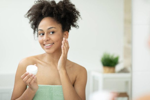Usmívající se Afroameričanka aplikující hydratační krém na obličej, zatímco drží sklenici v koupelně. Kvalitní fotografie - Fotografie, Obrázek
