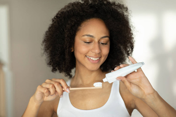 Uma mulher de pele escura sorri e espreme pasta de dentes em uma escova de madeira ecológica. Foto de alta qualidade - Foto, Imagem