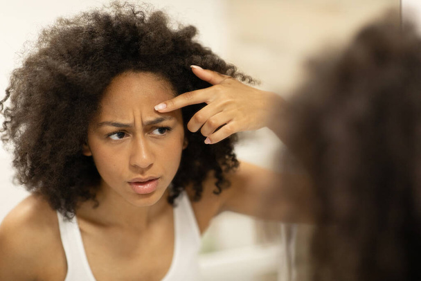 Unzufriedene schwarze Frau mit Problemhaut, die sich einen Pickel auf die Stirn drückt, steht in einem modernen Innenbadezimmer und leidet unter Akne. Hochwertiges Foto - Foto, Bild