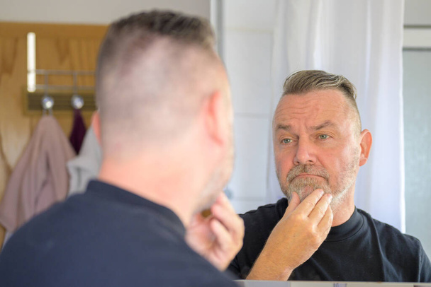Man checking out his neatly trimmed beard in the mirror running his fingers over the hair in an over the shoulder view - Photo, Image