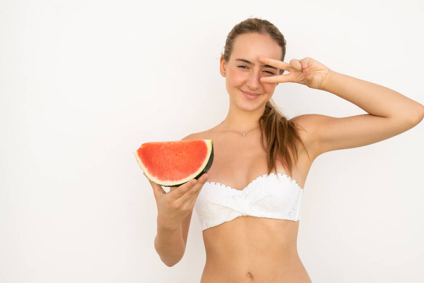 Jonge glimlachende vrouw in bikini die watermeloen eet en over een witte muur staat met vredessymbool. Summertime-concept. - Foto, afbeelding