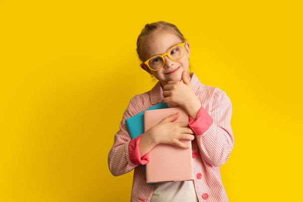 A girl with Down syndrome with textbooks is preparing to go to school, photo on a yellow background - Фото, изображение