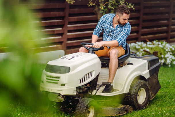 Professional gardener driving a riding lawn mower in a garden. Industrial landscaping work - Фото, изображение