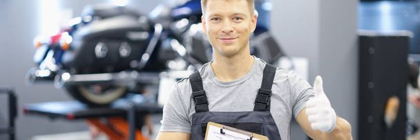 Porträt eines jungen Automechanikers, der Klemmbrett und Daumen in die Höhe hält. Empfehlungen für qualitativ hochwertige Wartung von Autos und Motorrädern - Foto, Bild