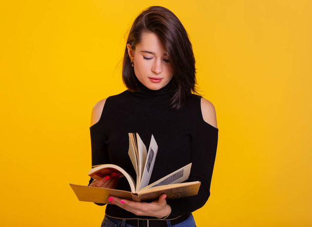 Pretty woman with a book in hands smiling at camera. Clever and excited lady holding open book. Studio shot isolated on yellow background - Фото, зображення