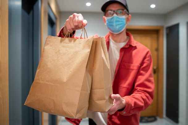 Delivery man in protective mask holding paper bag with food in the entrance. The courier gives the box with fresh vegetables and fruits to the customer. - Foto, imagen