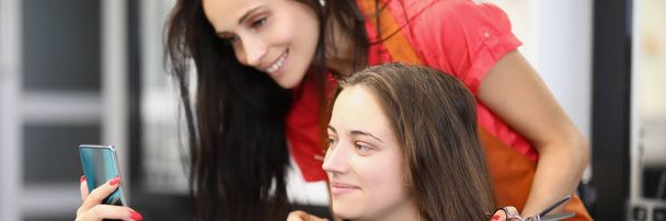 Portrait of smiling client on appointment at hairdresser. Woman show internet content to barber girl on smartphone. Change image, enter new life. Barbershop, style, beauty salon concept - Photo, Image