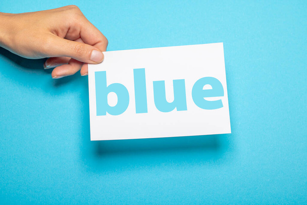 female hand holds a piece of paper with the word blue and copy space in front of a light blue background cardboard - Foto, imagen