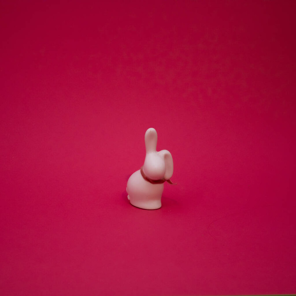 Ceramic figurine of a white rabbit with a decorative ribbon on a red background. Flat lay concept. - 写真・画像