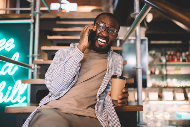Искренне черный мужчина в классических очках наслаждается кофе-брейком для создания позитивной сотовой связи через мобильное приложение - Фото, изображение