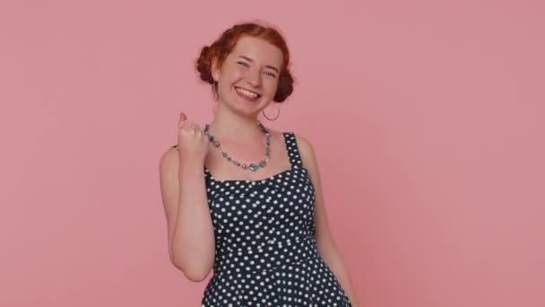 Happy redhead woman in polkadot dress shouting, raising fists in gesture I did it, celebrating success, winning, birthday, lottery goal achievemen. Young girl indoor on pink studio wall background - Filmati, video