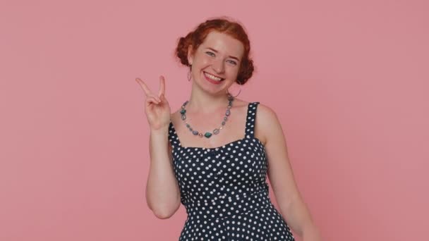 Die rothaarige junge Frau im Polkadotkleid zeigt das Siegeszeichen V, hofft auf Erfolg und Sieg, macht eine Friedensgeste und lächelt mit freundlich optimistischem Gesichtsausdruck. Ingwermädchen drinnen isoliert auf rosa Hintergrund - Filmmaterial, Video