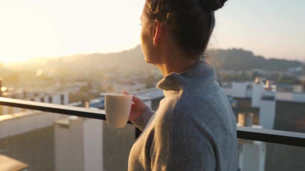 Kobieta zaczyna dzień od filiżanki herbaty lub kawy na balkonie o świcie, w zwolnionym tempie. Krajobraz miasta w tle. Nowoczesny miejski styl życia - Materiał filmowy, wideo