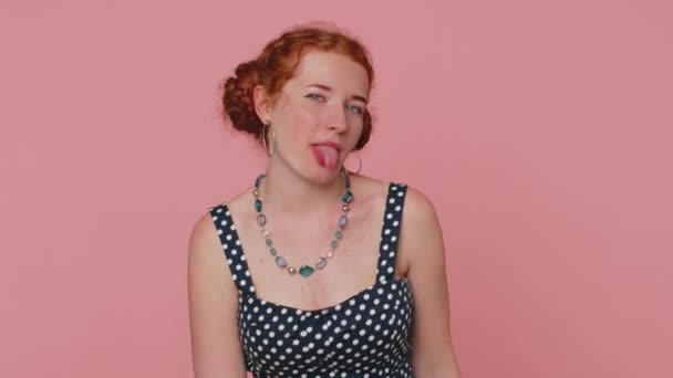 Cheerful funny young redhead woman showing tongue making faces at camera, fooling around, joking, aping with silly face, teasing. Ginger girl with freckles isolated alone on pink studio background - Metraje, vídeo