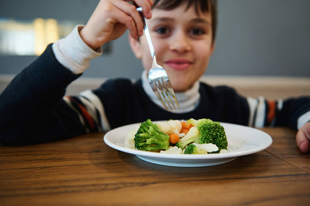 Focus sulle verdure al vapore, cibo vegano sano servito in piatto bianco e ragazzo adorabile offuscata tenendo la forchetta e sorridente guardando la fotocamera durante il pranzo in mensa - Foto, immagini