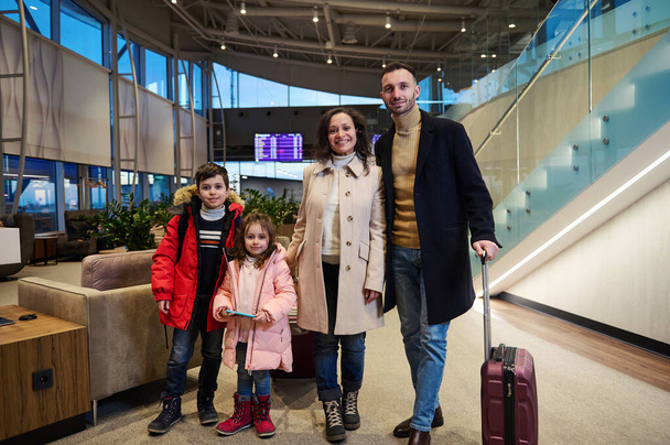 Cheerful family with suitcases, against flight information board with timetable in international airport departures terminal. Air travel concept, family relationships, children and parents together - Фото, изображение