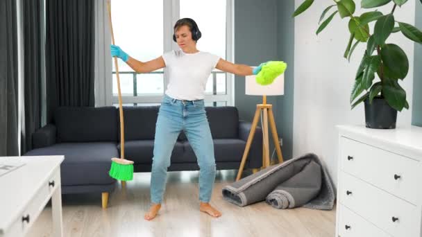 Blanke vrouw in koptelefoon schoonmaken van het huis en plezier hebben dansen met een bezem en washandje - Video
