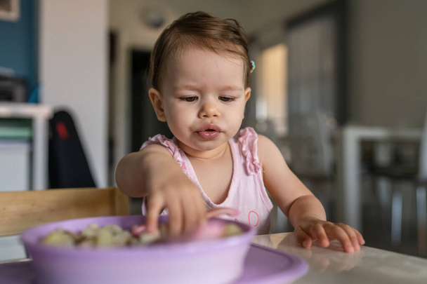 One child small caucasian toddler female baby eating at the table alone at home in room real people copy space early child development concept learning front view - Zdjęcie, obraz