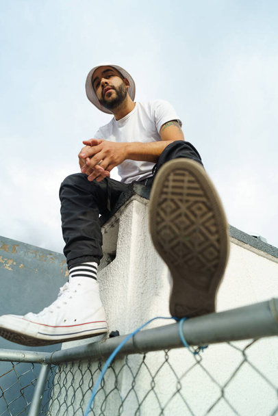 Latin young man with black hair and beard wears comfortable and casual clothes sitting with heaven in the background, lifestyle and urban fashion, male model - 写真・画像