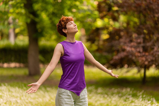 Mulher lésbica calma em roupas casuais espalhando braços e fechando os olhos enquanto respira ar fresco no dia de fim de semana no parque - Foto, Imagem